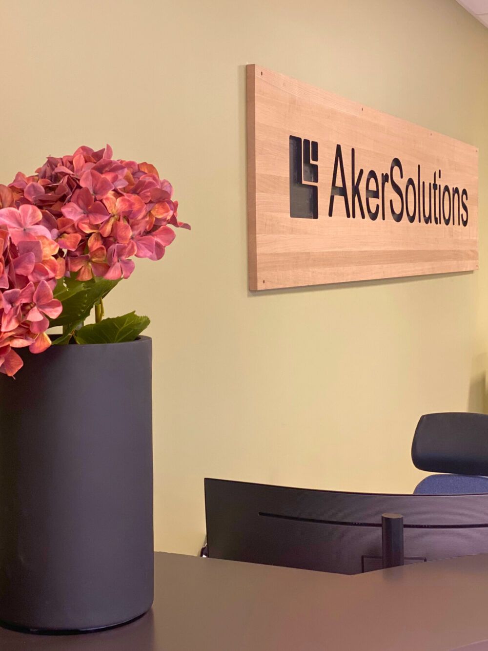 Aker Solutions sin logo inngravert i tre på vegg bak resepsjonen på Akso. Svarte detaljer på skranken med blomster som frisker opp miljøet. Kontorstol i blått ullstoff.